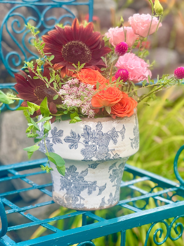 A decorative antique white ceramic flower pot with a slate gray floral pattern, filled with a colorful bouquet of flowers including orange roses, pink lisianthis, chocolate sunflowers, and green foliage, displayed on a teal metal garden bench.